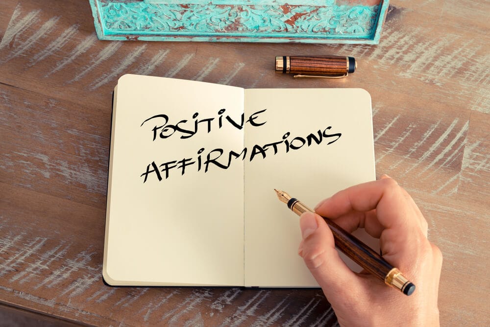 A person with a fountain pen in their hand writes "positive affirmations" in their journal.