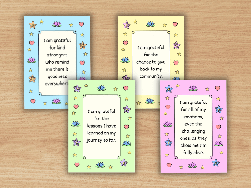 Four affirmation cards on a wooden table. The four affirmations are: "I am grateful for kind strangers who remind me there is goodness everywhere," "I am grateful for the chance to give back to my community," "I am grateful for the lessons I have learned on my journey so far," and "I am grateful for all of my emotions, even the challenging ones, as they show me I'm fully alive."