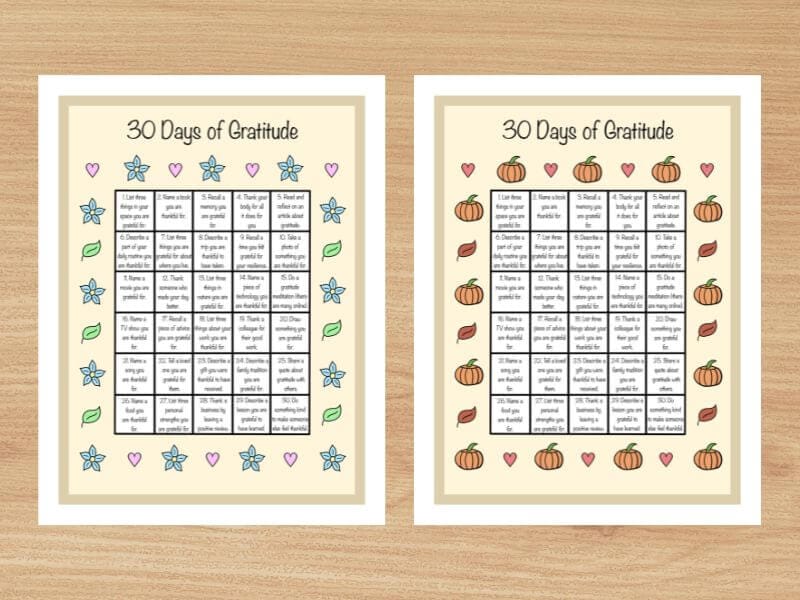 Two pieces of paper side by side on a desk. They are each titled "30 Days of Gratitude" and contain a grid in the middle of the page filled in with 30 gratitude prompts. The border around the grid of the first piece of paper is floral and made of blue flowers, pink hearts, and green leaves. The border around the grid on the second piece of paper is fall themed, with orange pumpkins, red hearts, and brown leaves.