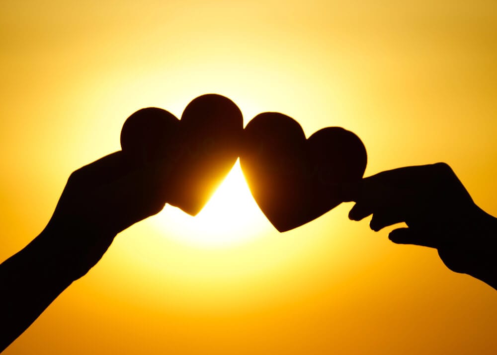 Two hands holding hearts together in front of the Sun.