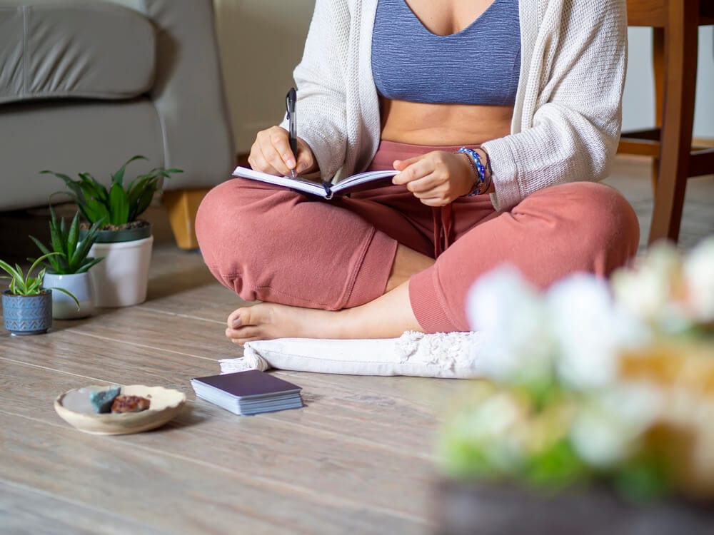 A woman sits cross-legged on the floor, writing in a journal. In front of her are crystals and oracle cards.