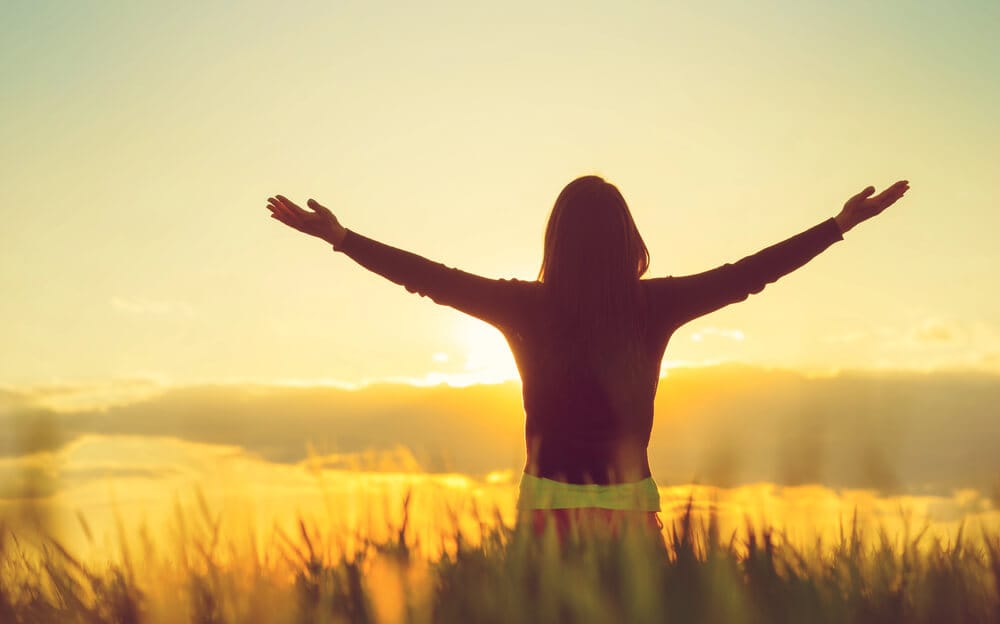 A woman stands with her arms outstretched and her back to the camera, watching the sun rise.